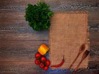 On the wooden table is for table linen cloth, a wooden spoon and fork, yellow peppers bunch of parsley and cherry tomatoes on a branch