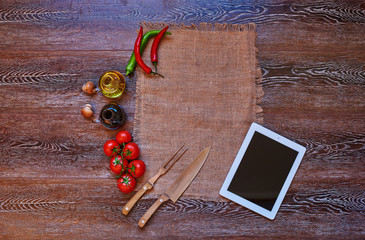 On the kitchen linen towels on a wooden table lies textural tablet, next to the digital  tablet are vintage silver knife and fork, tomatoes and hot peppers for cooking meals