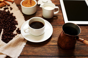 Coffee background. White tablet computer blank black screen with white cup of coffee and coffee beans on dark wooden background. Freshly made coffee in old rustic desk. Top view with copy space..