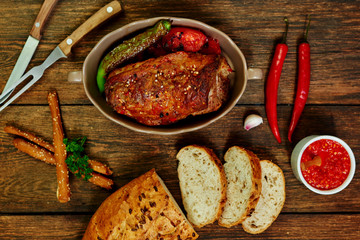 Baked pork loin  in ceramic form with fork, knife and homemade rustic bread on dark rustic background. Roasted pork served with grilled vegetables and tomato sauce. Cozy village dinner..