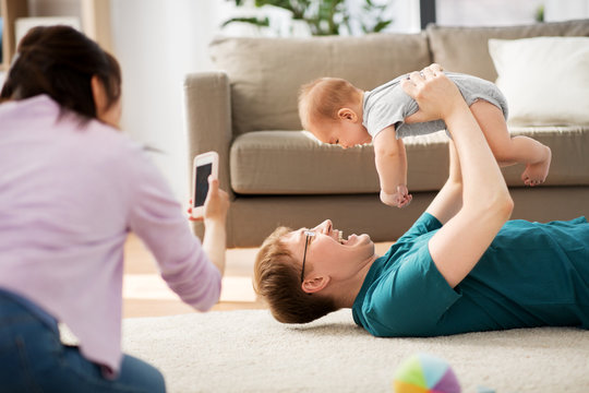 family, technology and people concept - happy mother and father taking picture or recording video of baby boy with smartphone at home