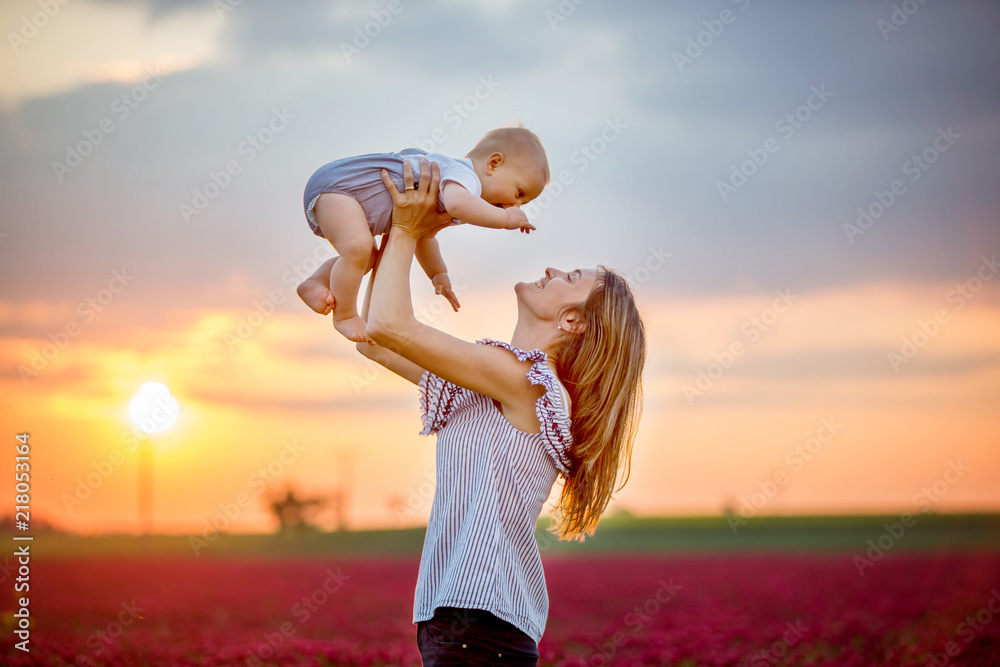 Sticker Young mother, embracing with tenderness and care her toddler baby boy in crimson clover field, smiling happily