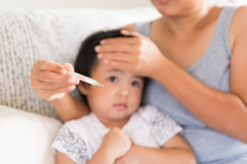 Close up of a mother checking the temperature of her ill baby with a thermometer on a couch in the living room at home. Medicine and health care concept.