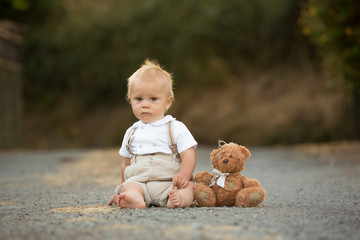 Handsome stylish toddler child boy with funny face in suspenders playing in spring or summer garden or park with his teddy bear
