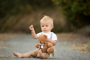 Handsome stylish toddler child boy with funny face in suspenders playing in spring or summer garden or park with his teddy bear
