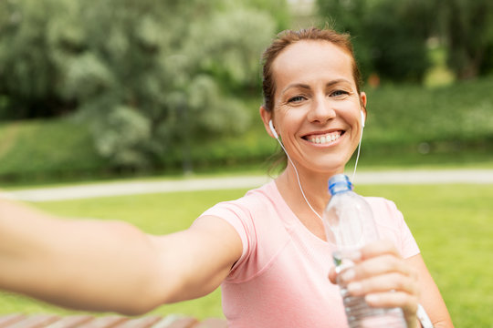 fitness, sport and healthy lifestyle concept - happy woman in earphones and water taking selfie in park