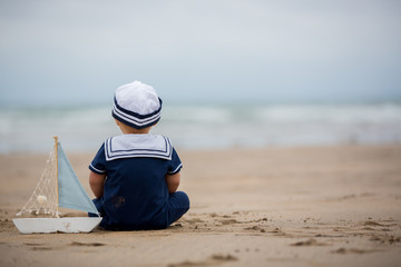 Baby boy sitting on the beach near the water and plays with a toy ship and bear toy