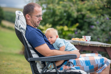 Young father, holding his cute baby boy outdoor, cuddle him, kissing and huging, sitting outdoor on the porch