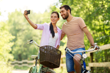 people, technology and lifestyle concept - happy couple with bicycles taking selfie by smartphone at summer park