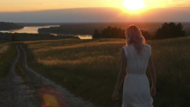young romantic woman walking on the road in the field at sunset