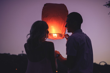 silhouettes of couple launching sky lantern on river beach in evening and looking at each other - obrazy, fototapety, plakaty