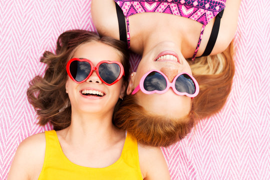 summer fashion, leisure and valentines day concept - smiling teenage girls in heart shaped sunglasses lying on picnic blanket