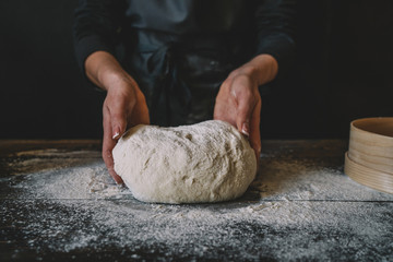 Woman chef in a home kitchen prepares the dough with flour to make the Italian pasta. The concept of nature, food and diet. Ball of pizza dough on a rustic wooden background with dusting of flour.