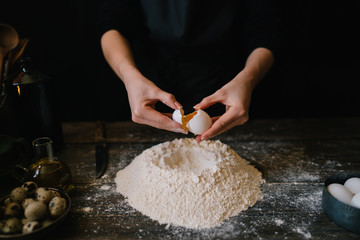 Egg poured into flour in a rustic wooden table during the baking process. Baking ingredients on the dark wooden table. Making bread, retro styled imagery. Grain added. Selective focus.