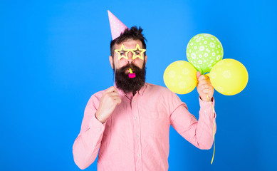 Man with beard on calm face holds air balloons, blue background. Surprise concept. Guy in party hat...