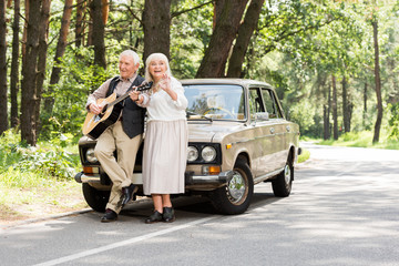 senior wife with husband playing guitar leaning on car on road in forest