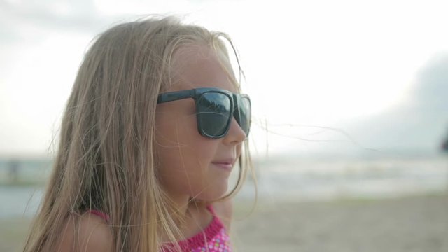 Little beautiful daughter with long hair and a pink swimsuit sitting on the beach in sunglasses, on the background of the sea