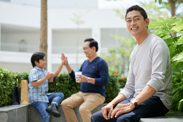 Handsome Asian guy smiling and looking at camera while sitting on border in park near high-fiving father and son