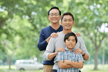 Three male members of Asian family smiling and looking at camera while standing on blurred background of park