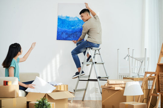 Young Asian lady showing boyfriend where to hang nice picture while decorating new apartment together