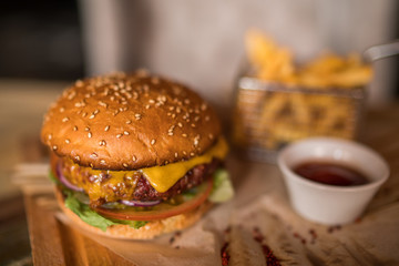 Delicious fresh homemade burger with spicy tomato sauce and beef on wooden background. Tasty homemade grilled beef burger with lettuce, cheese and onion served on cutting board on a brown wooden table