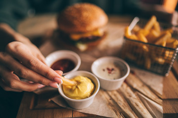 Woman's hand dipping french fries in barbecue sauce. Delicious fresh burger with tomato and cheese. Fast food concept. Beef burger in hands with french fries on rustic wooden background.