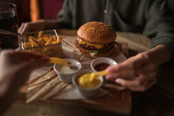 Woman's hand dipping french fries in barbecue sauce. Delicious fresh burger with tomato and cheese. Fast food concept. Beef burger in hands with french fries on rustic wooden background. Soft focus.