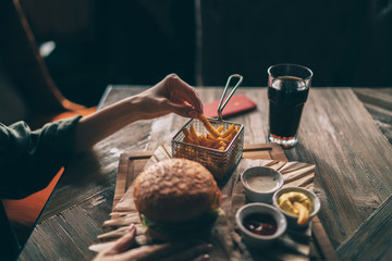 Woman's hand dipping french fries in barbecue sauce. Delicious fresh burger with tomato and cheese. Fast food concept. Beef burger in hands with french fries on rustic wooden background.