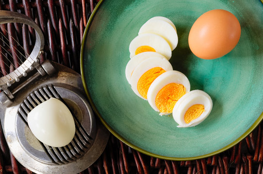 Hard-boiled egg cut and piled on egg slicer and full egg with shell on green plate or dish.