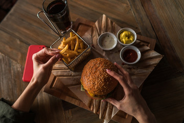 Woman's hand dipping french fries in barbecue sauce. Delicious fresh burger with tomato and cheese. Fast food concept. Beef burger in hands with french fries on rustic wooden background.