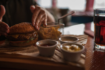 Woman holds burger with hands and fries on the background in cafe. Fresh burger cooked at barbecue in craft paper. American food. Big hamburger with meat and vegetables closeup unfocused at background