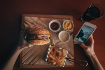 Women's hand using an application to send an sms message in her smartphone device while eating. People and eating concept. Hamburger, french fries, ketchup, mustard on a wooden board. Toned image.