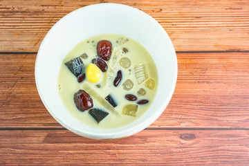 Fototapeta na wymiar soybean milk extra ingredients with black grass jelly, ginkgo, longan, red bean, pivot seed ,thai local famous street food in white bowl on wooden table background