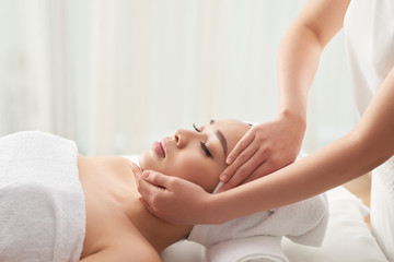 Obraz na płótnie Canvas Crop hands of massage therapist rubbing face of pretty female client while working in spa salon