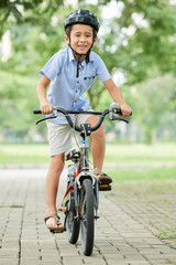 Cute Asian boy in helmet smiling and looking at camera while riding bicycle in park on sunny day
