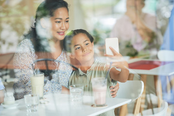 Asian woman and cute girl using smartphone to take selfie while sitting behind window in cafe
