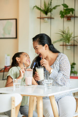 Adult Asian woman and adorable girl laughing and looking at each other while drinking delicious milkshakes in cozy cafe