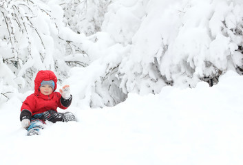 Fototapeta na wymiar Cute toddler boy in winter suit sitting on a big snowdrift and raises his hand in greeting, surrounded by snow-covered trees. Snowy winter. Cope space