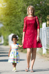 A white woman is walking along the street with a black girl. A European mother with an African daughter in summer dresses is walking with her hand.