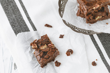 Homemade Brownie with Pecans on Parchment Paper; Brownies Stacked on Pedestal in Background; Black & White Kitchen Towel