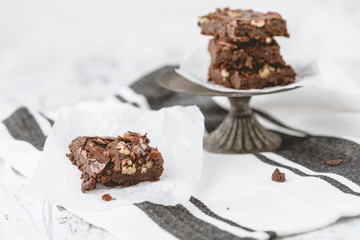 Homemade Brownie with Pecans on Parchment Paper; Brownies Stacked on Pedestal in Background; Black & White Kitchen Towel