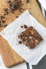 Single Homemade Brownie with Pecans on Cutting Board with Semi-Sweet Chocolate Chips, Unsweetened Chocolate, and Cocoa Scattered Around; Black & White Kitchen Towel in Background