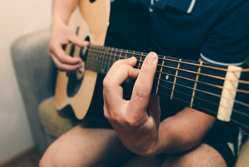 Obraz premium Man at home is playing the guitar. Guys hands are taking the chord on strings. Music making lifestyle concept. Free time hobby for everyone. Retro guitar photo.