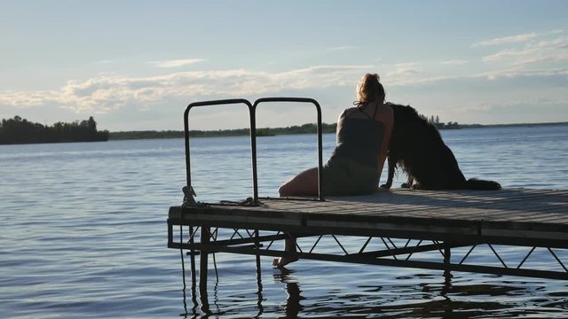 Woman sits at the end of a dock with her dog. She pets her dog and looks at the water.