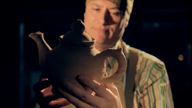 A craftsman at a pottery checks a clay kettle, then looks at a camera.