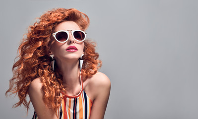 Fashion Portrait Sensual Redhead Girl with Trendy Wavy Curly Hairstyle in Studio. Beautiful Attractive Model woman in Stylish Summer Sunglasses. Close up female Face.