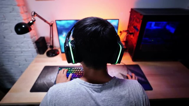 Back view of asian male gamer playing computer game by the table at home