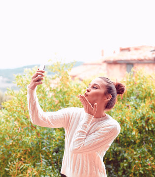 Beautiful young woman in white sweater and underwear, sending kiss to phone, standing on balcony. Tuscany, Italy. Small depth of field.