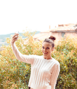 Beautiful young woman in white sweater and underwear, taking selfie, standing on balcony. Tuscany, Italy. Small depth of field.