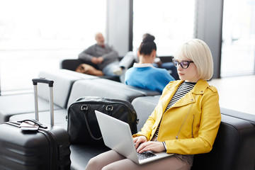 Young attractive businesswoman sitting with luggage and typing on laptop in hall of airport